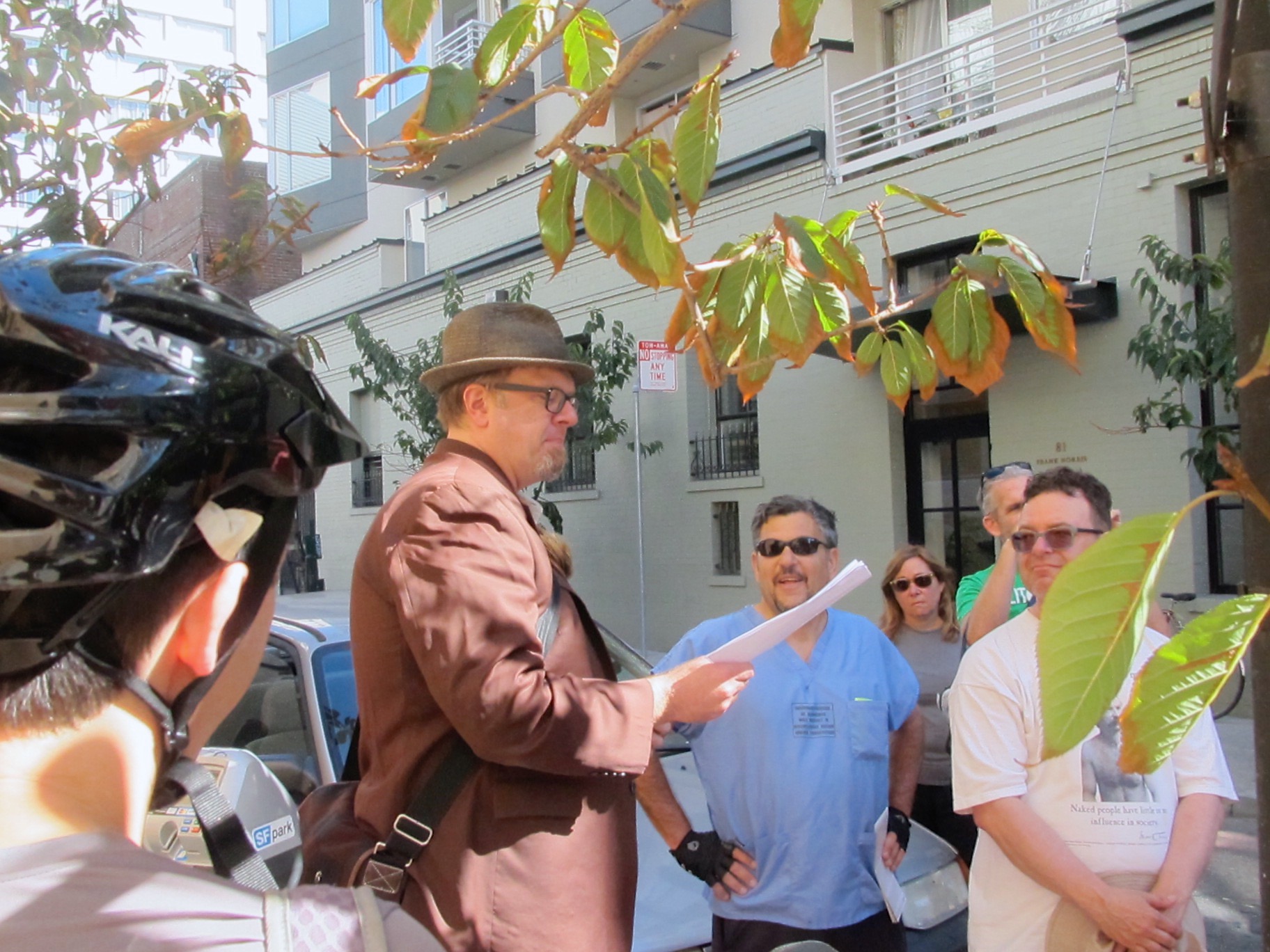 A reader, Jim Nelson, wearing a brown hat and suit jacket, reads to a group of bicycle riders on an urban side street in San Francisco.