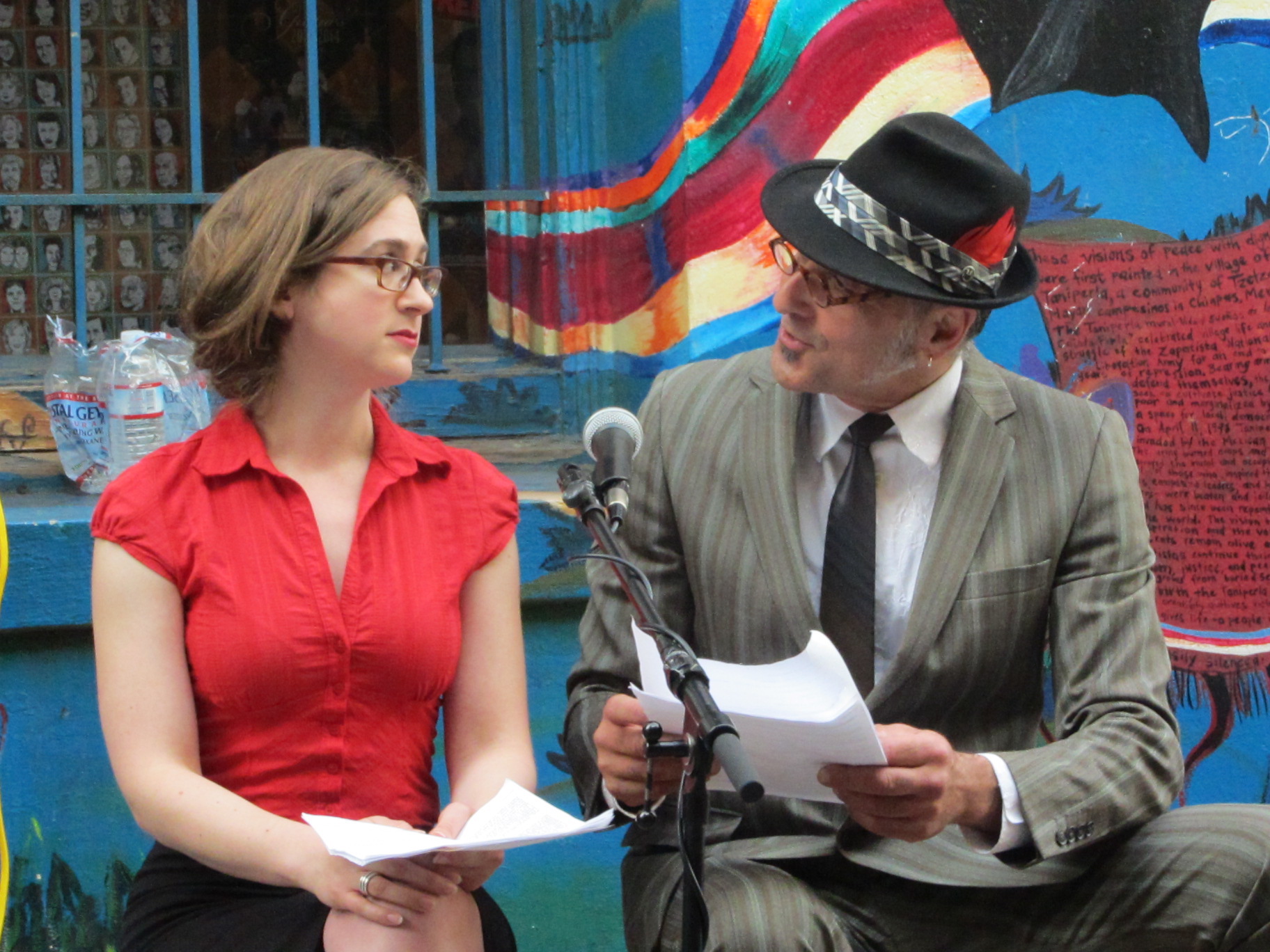 Two performers, Beck Hirshfeld, a young woman in a red button down shirt and glasses, Steffanos X, a middle-aged man in a grey striped suit and a nice fedora with a red feather, seated in front of a colorful mural.