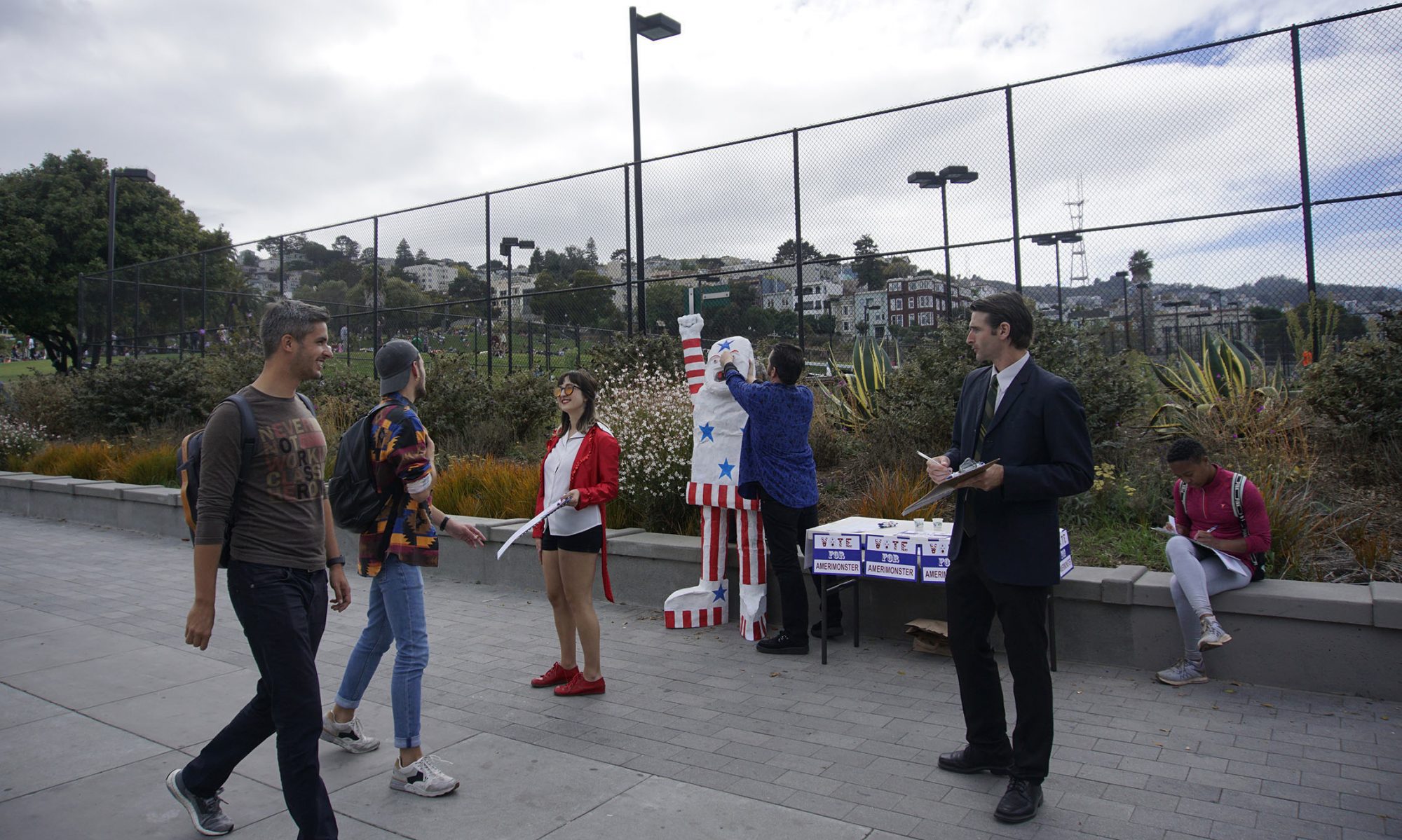 Small group of costumed performers registering voters on sidewalk. A large American Flag Golem, the Amerimonster, stands in the background with an upraised fist.