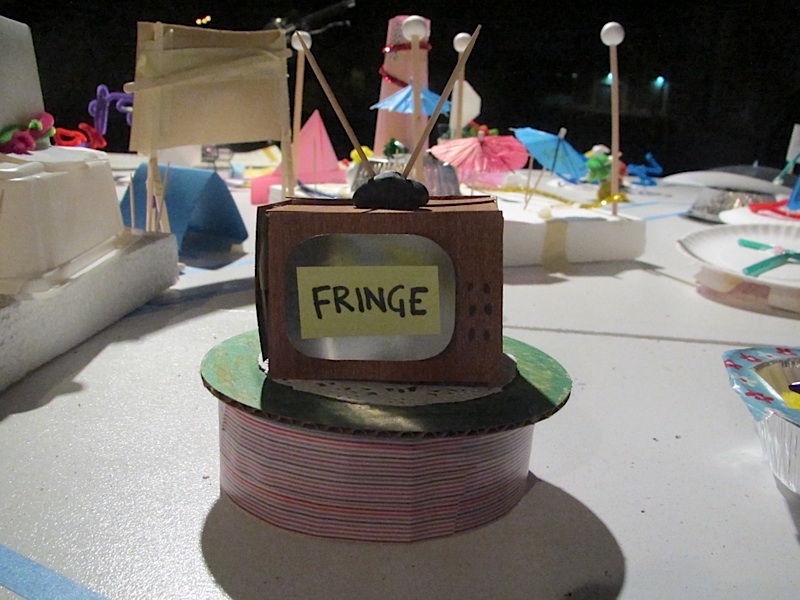 A craft table with a cardboard television on top of a ribbon reel with Fringe written on the front