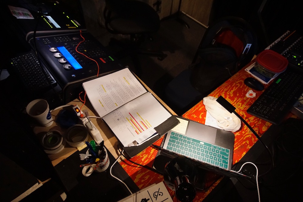 A low wooden shelf at right angles to a tech table, both covered with computers, lightboard, tech script, and coffee cups.
