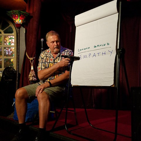 Performer Ed Wolf, wearing a shirt patterned with Loteria game pieces and green shorts points a gun at a large sheet of paper on an easel, with the words "common ground; empathy" written on it.