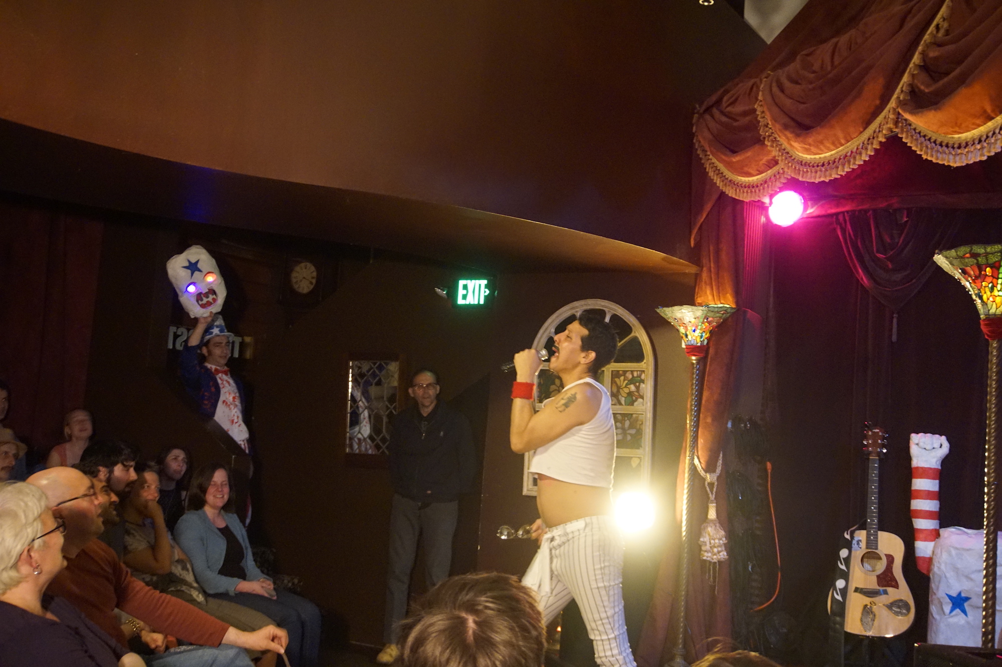 Performer Peter Griggs dressed like Freddie Mercury in a white crop top, sings into a microphone in front of a smiling crowd.