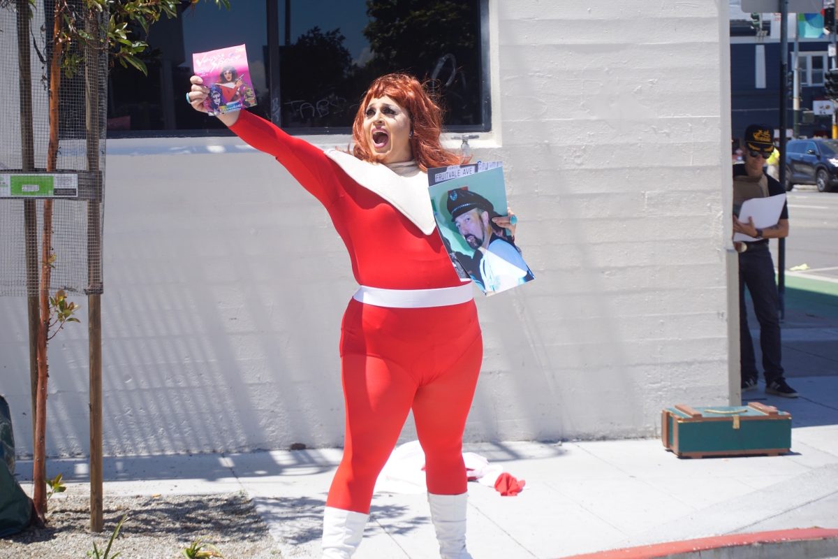 Performer Landa Lakes, a drag queen in a red and white spandex space suit, holds up flyers and photographs on a corner of Ringold Alley in San Francisco.