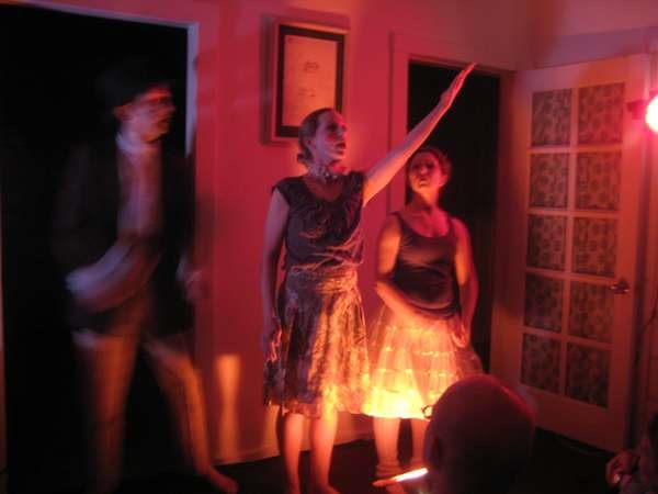 Three Dancers stand in a living room doorway, dim red lighting and artistically blurred image.