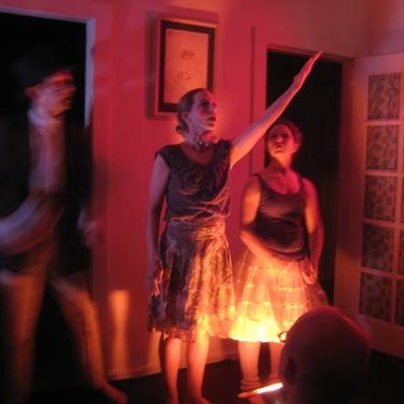 Three Dancers stand in a living room doorway, dim red lighting and artistically blurred image.