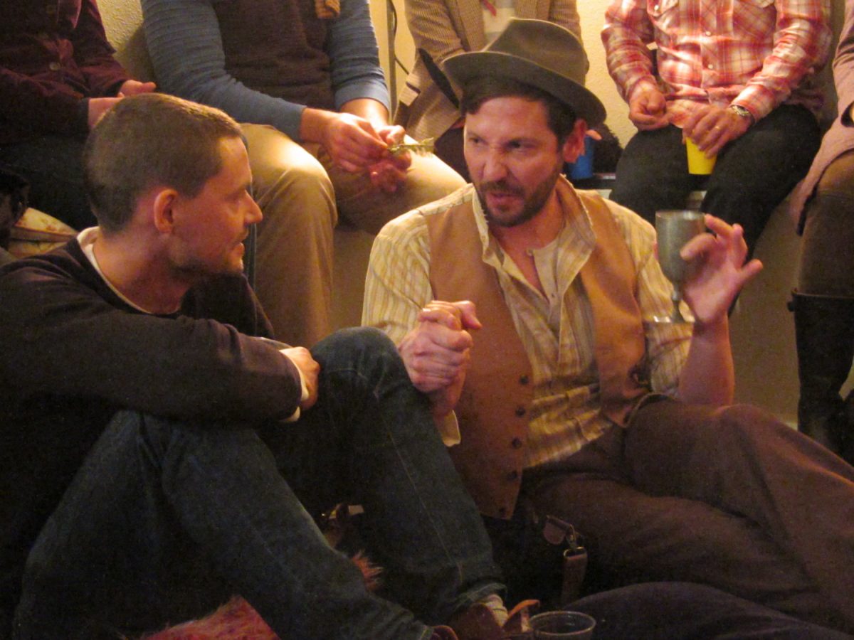 Performer Ryan Hayes in tan-colored vest, brown brimmed hat and yellow-striped shirt sits on the floor next to an audience member in a dark pullover top while holding a metal wine glass.