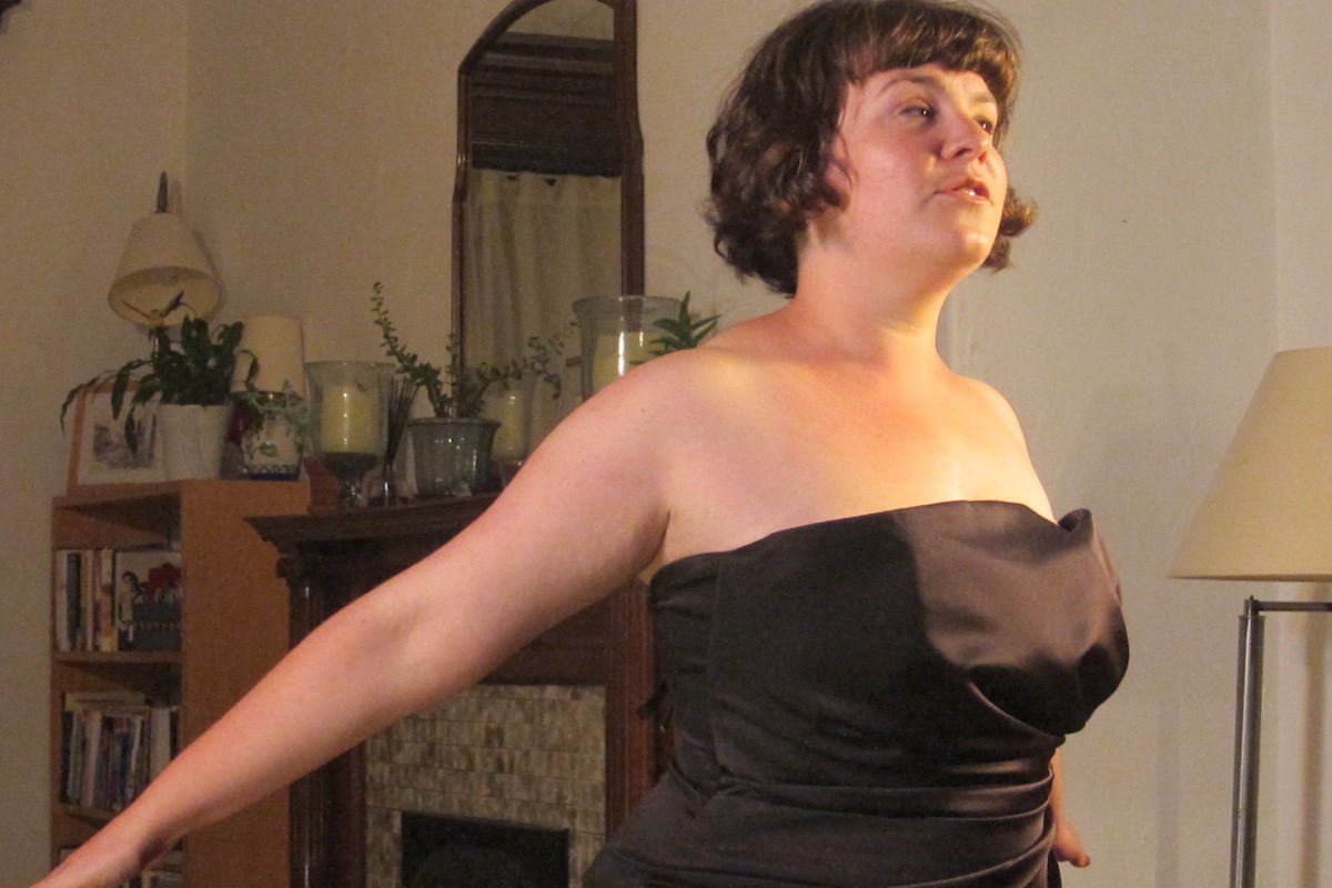 A performer in a black cocktail dress and short dark hair stands in a living room, arms outstretched behind her.