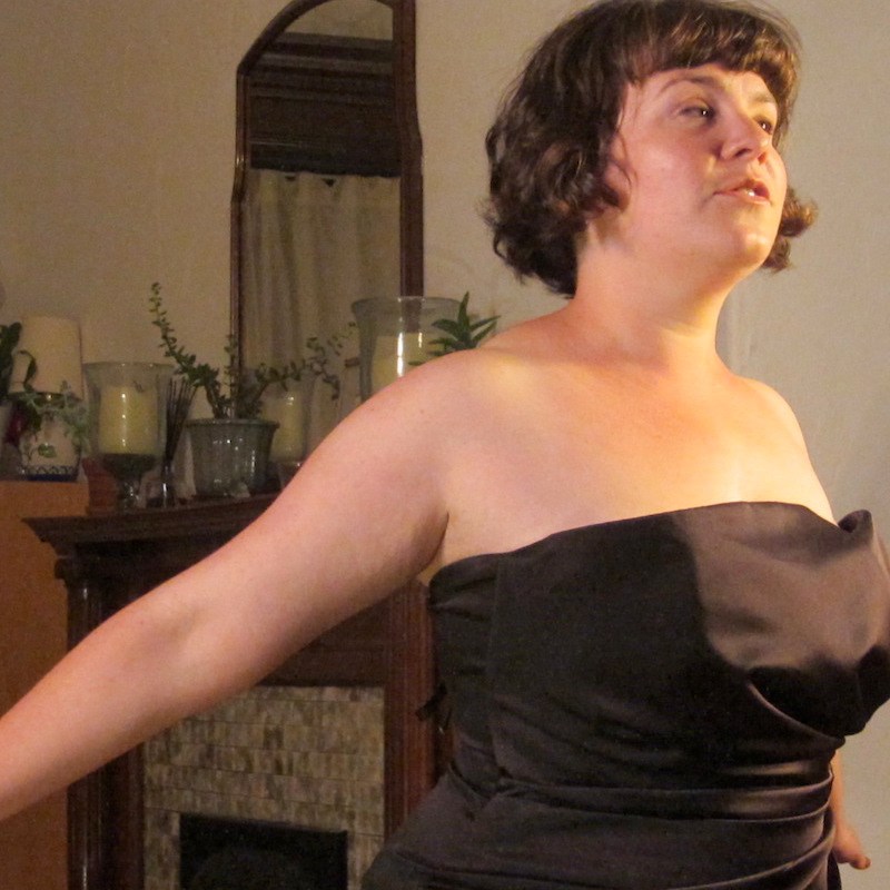 A performer in a black cocktail dress and short dark hair stands in a living room, arms outstretched behind her.