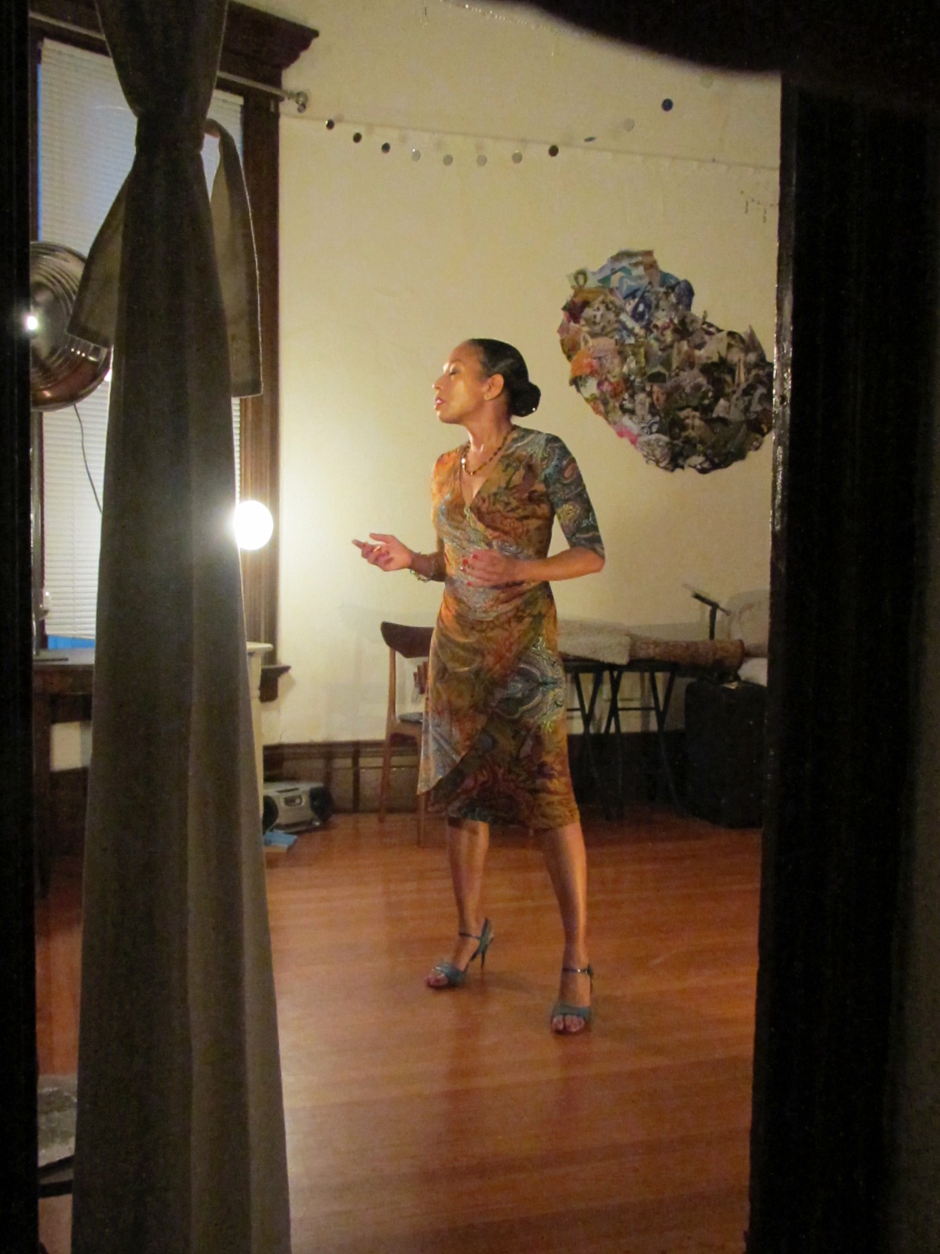 Performer Mia Paschal, a Black Woman with pulled back dark hair, stands in a living room on a wood floor in high heeled tango shoes and a wraparound dress of patterned grey and brown.