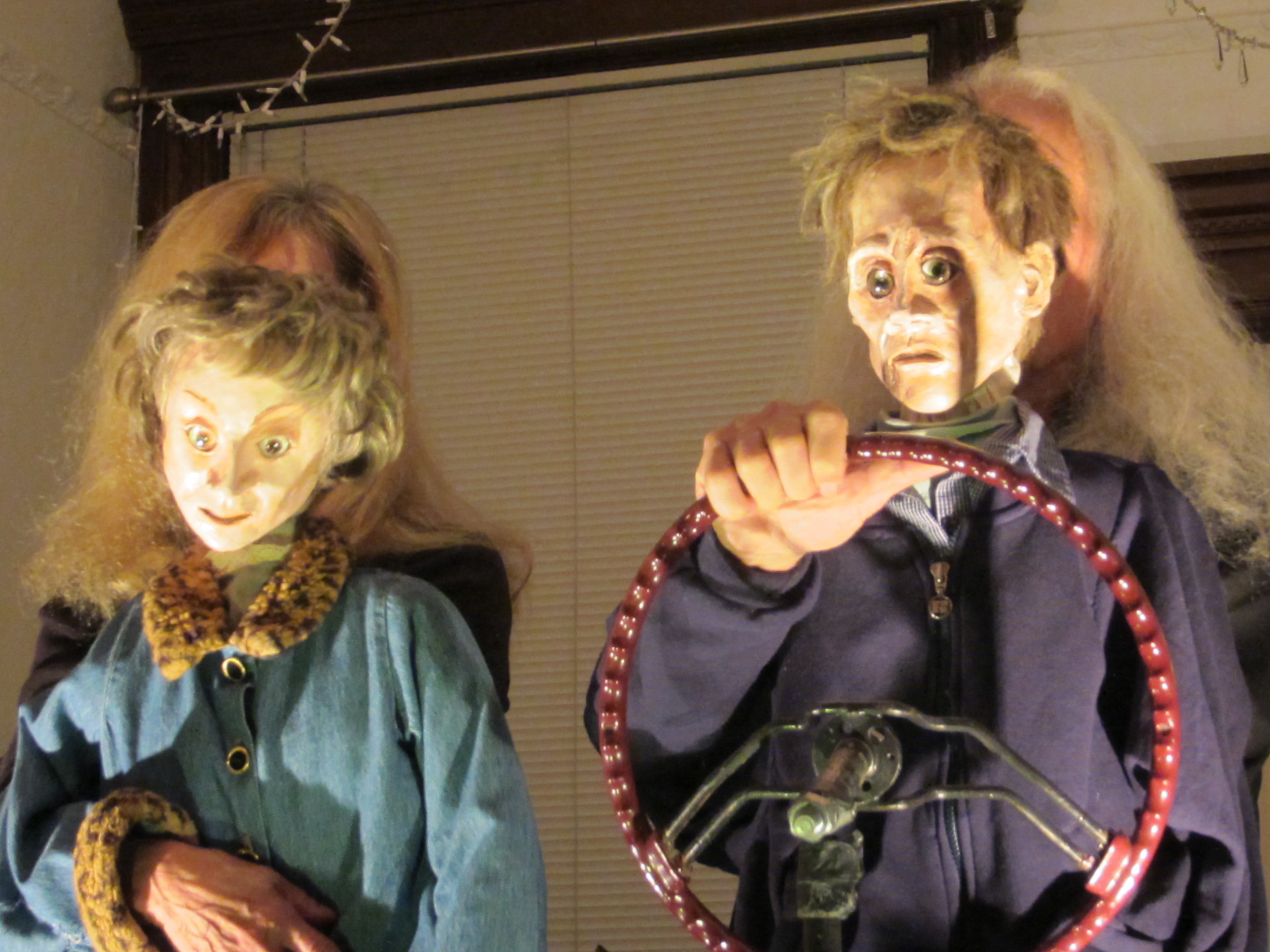 Two elderly performers wearing human-face masks pretend to drive a car in a private living room.