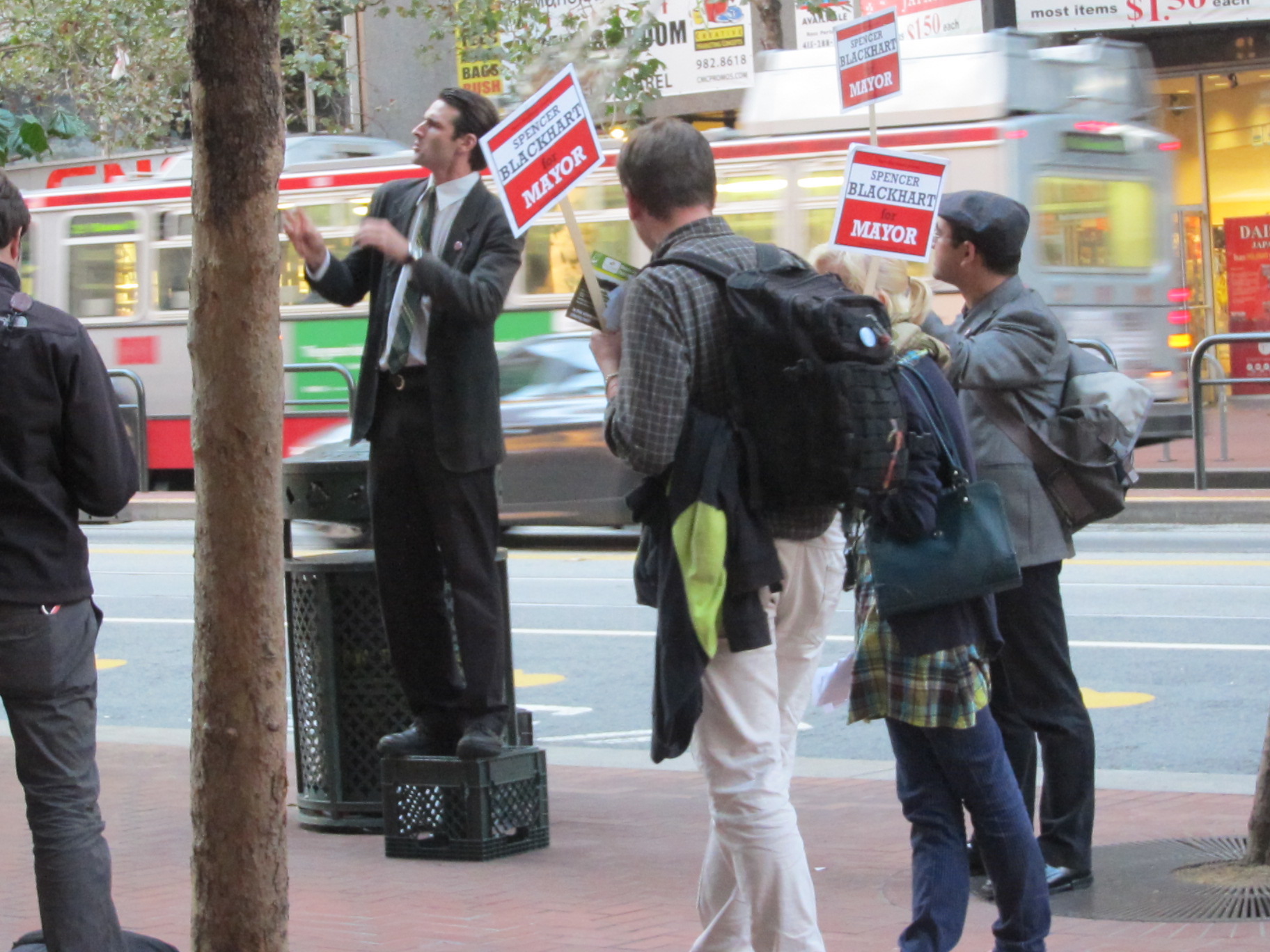 Spencer Blackhart performer in a dark suit and striped tie stands on a milk crate on a city sidewalk, as people carrying Spencer Blackhart for Mayor signs surround him.