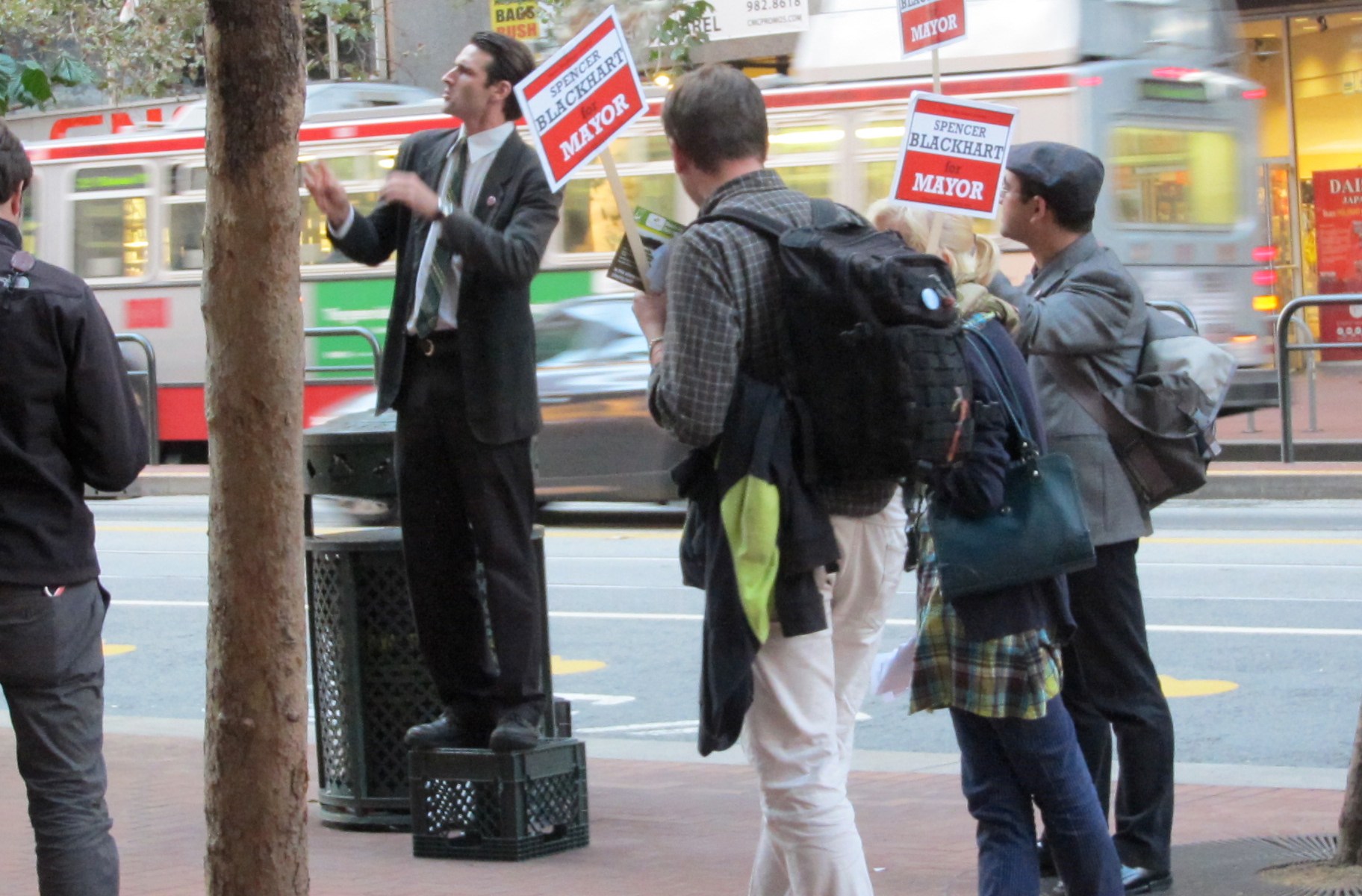 Performer Spencer Blackhart in a suit, standing on a milk crate on a busy sidewalk, surrounded by supporters carrying Spencer Blackhart for Mayor signs.