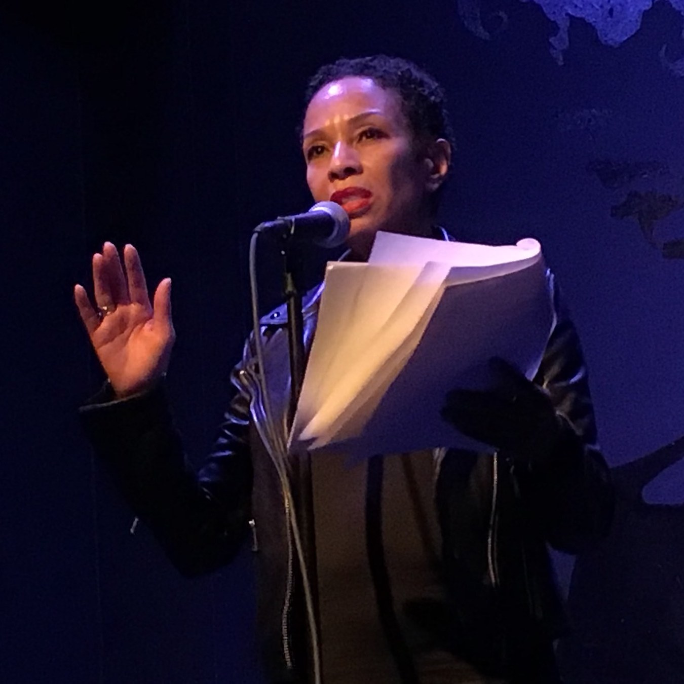 Performer Mia Paschal in a black leather jacket reads from a script onstage.