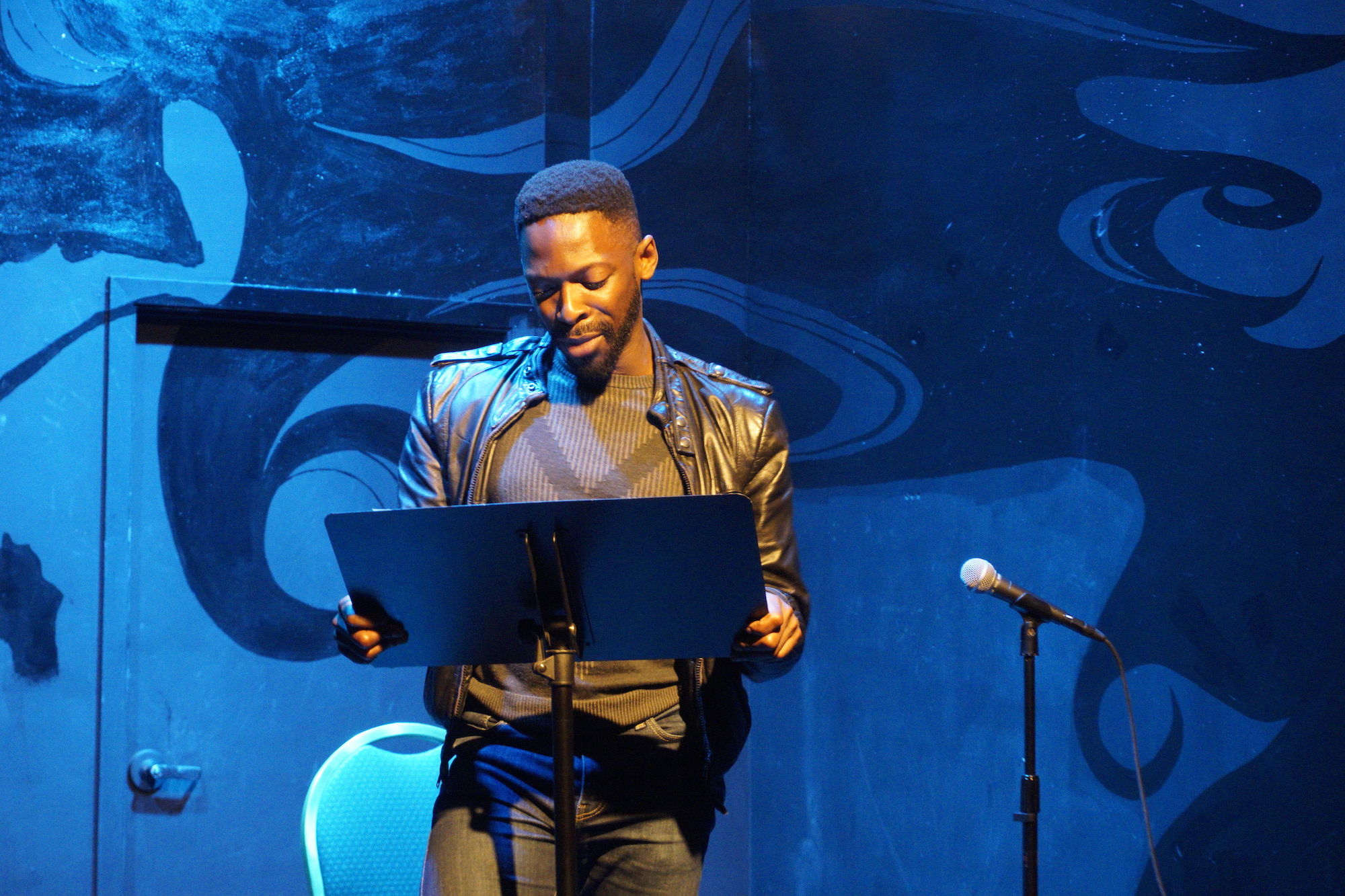 Performer Rotimi Agbabiaka in a black leather jacket and blue lighting stands behind a music stand onstage.