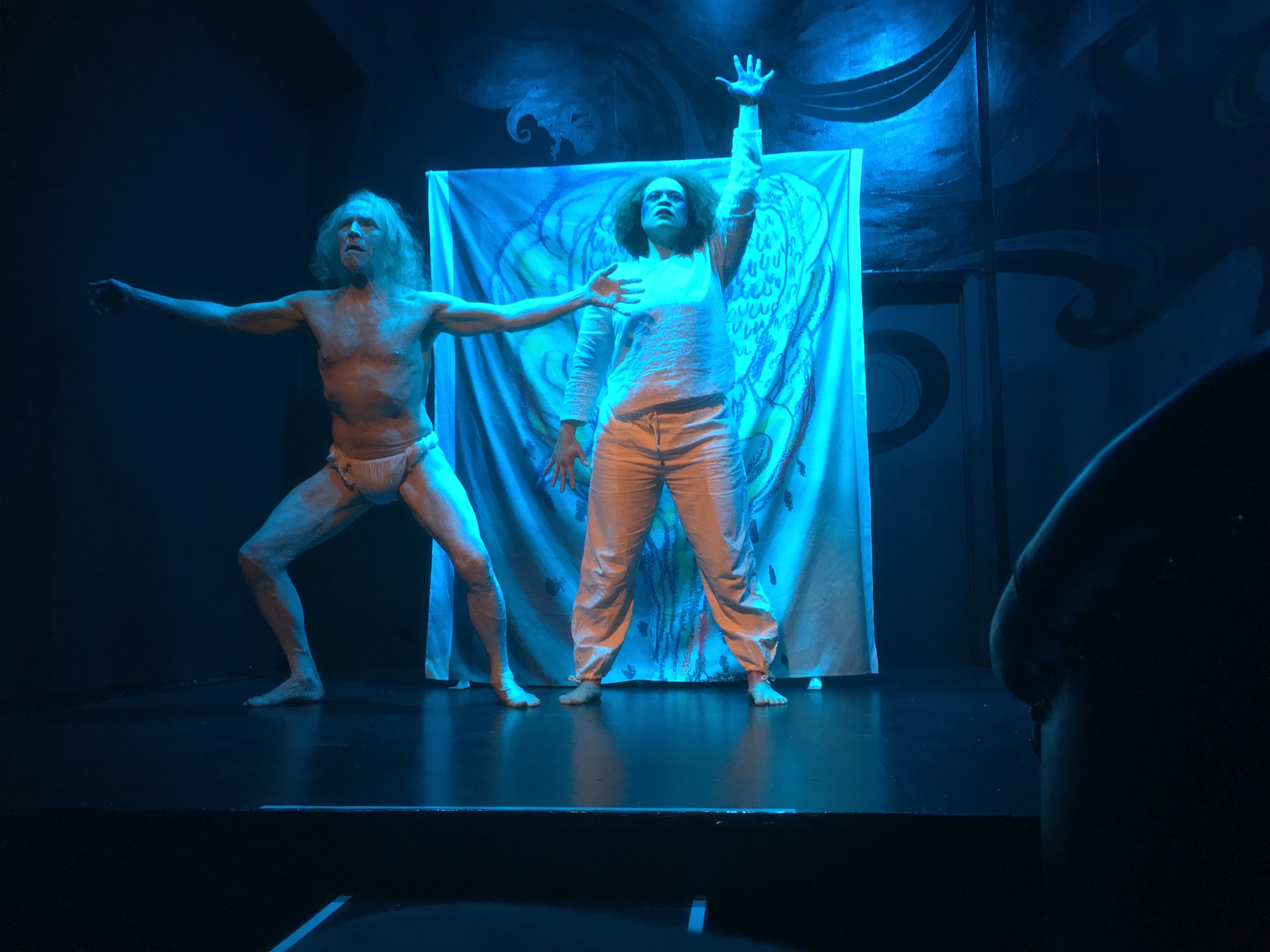 Two dancers, Eric Larsen, an elderly male in loincloth and Ashton Wolgast, a Black woman in loose dance clothing, perform in front of a backdrop painted with a mushroom cloud..