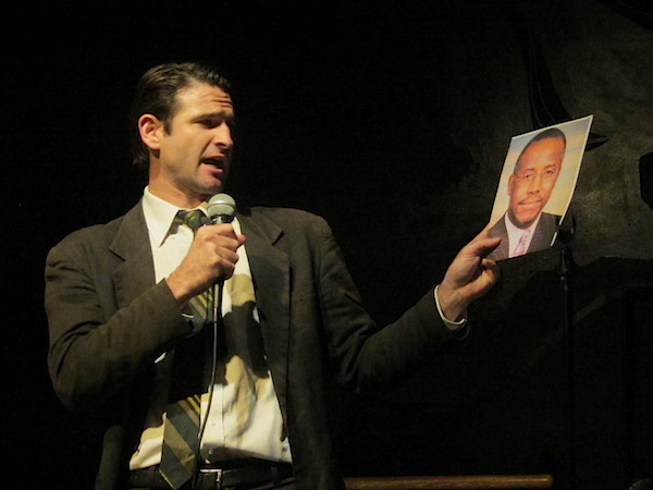 Performer Spencer Blackhart dressed in a dark suit and striped tie, holding a microphone and a photograph of a Black politician.