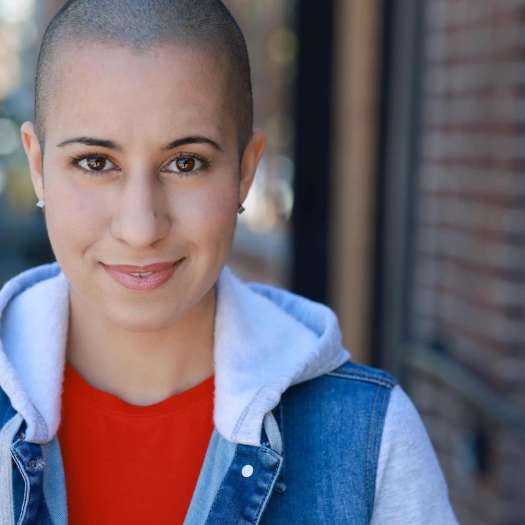 Edna, a young woman with a shaved head and brown eyes, earing a red t-short and denim jacket, smiling at the camera