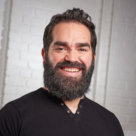 Nathaniel Justiniano, a man with dark hair, a full beard with some grey hairs in it, and brown eyes, wearing a black cotton shirt and smiling at the camera