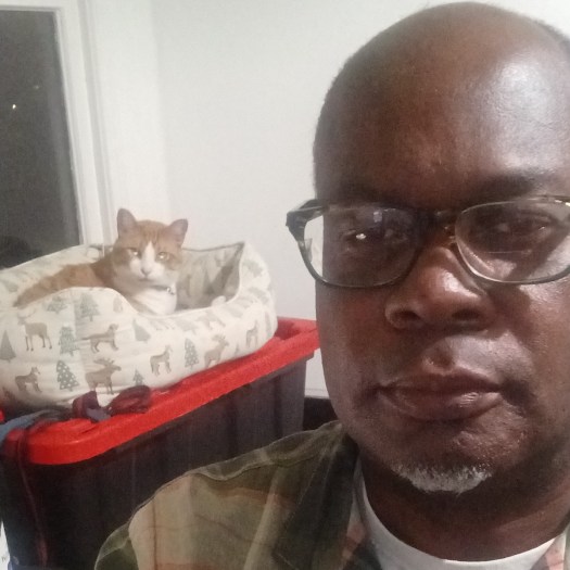 Patrick Simms, a Black man with a grey goatee looks at the camera as a cat in a cat bed watches from behind him