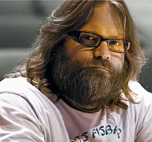 Cliff Caruthers, a white man with long brown hair and a beard, wearing thick, black-rimmed glasses and a white sweatshirt, looks at the camera