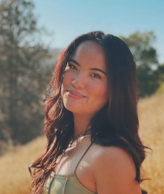 Jerikka Gamboa, a young woman with long dark hair and a medium complexion stands outdoors in a sunny field and smiles at the camera