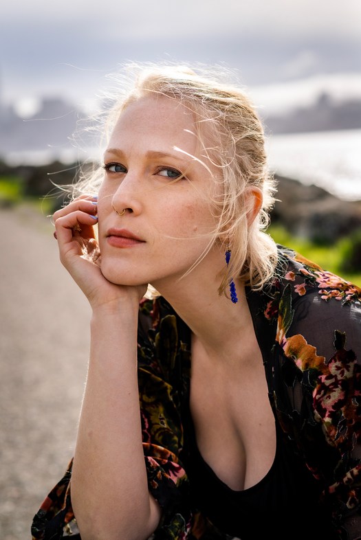 Megan Trout, a young woman with blond hair, wearing a dark floral top and blue dangling earrings, in three-quarter profile with her hand on her face.