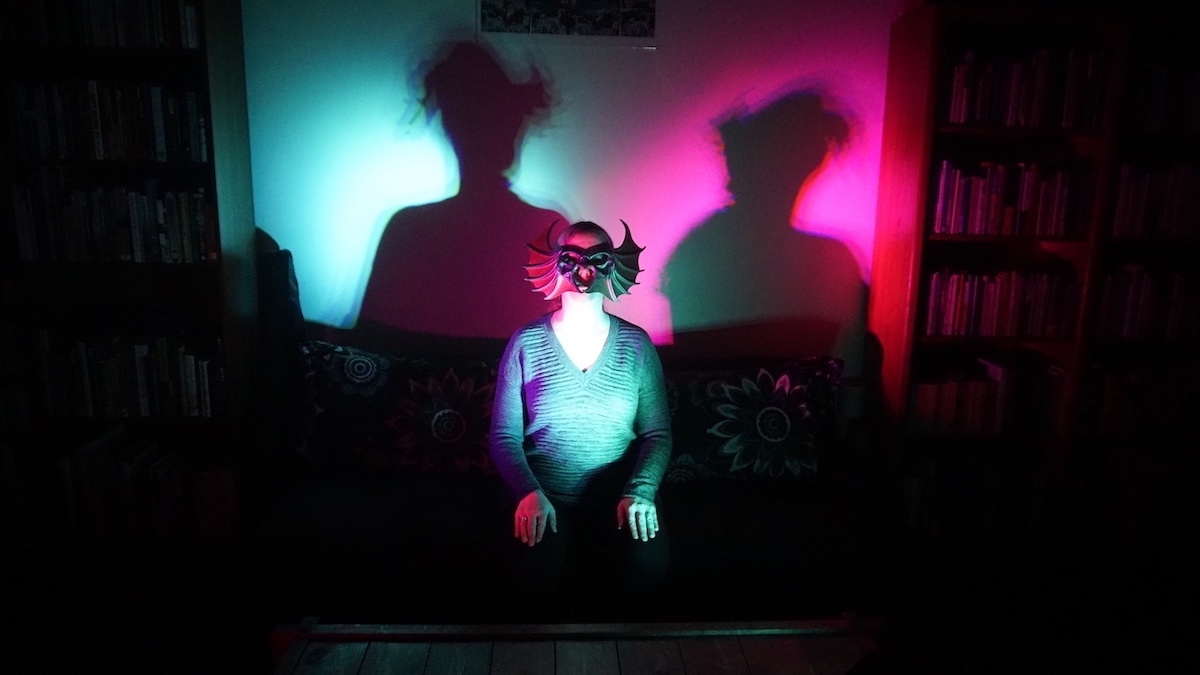 A person in a monster mask sitting in a dark room with a teal colored light and a magenta light shining on them from the side angles