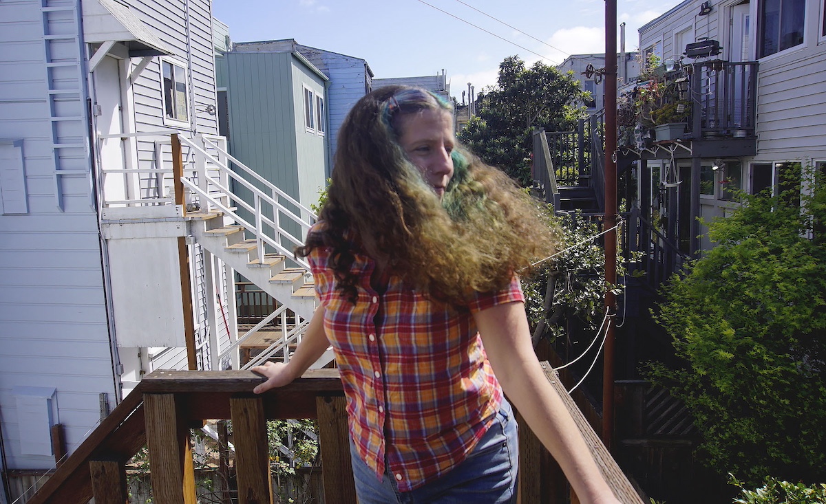 A woman standing outside on a deck, with apartment buildings and backyard gardens behind her. She has dark wavy hair with a green streak and is wearing an orange plaid shirt.